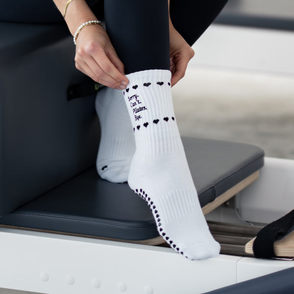 Weiße Pilates Grip Socks mit schwarzen Herz-Details und dem Spruch ‚Sorry. Can’t. Pilates. Bye.‘ auf einem Reformer – stylische rutschfeste Socken für effektives und sicheres Pilates-Training
